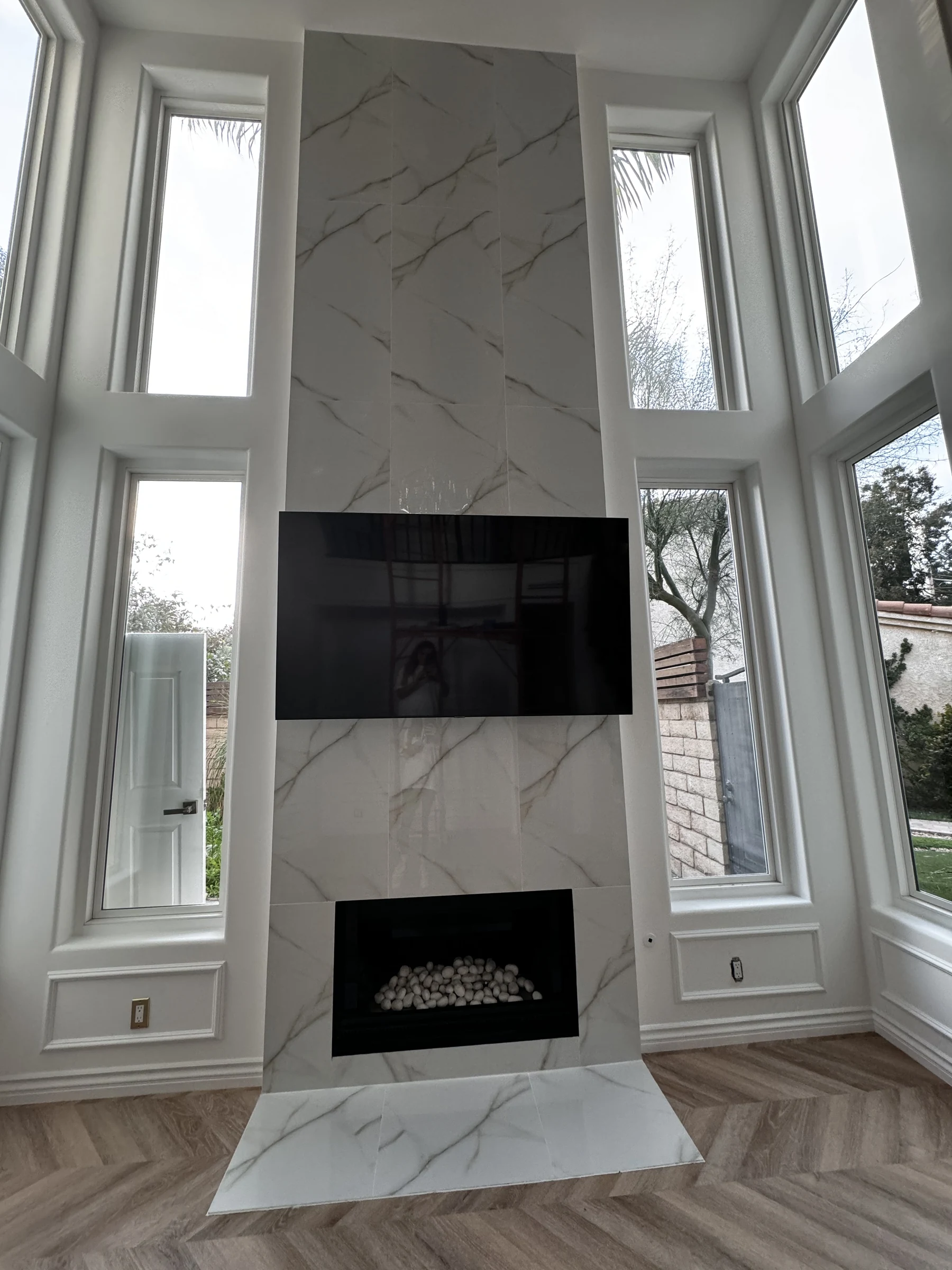 Marble fireplace wall between tall windows over a herringbone floor in Newport Beach